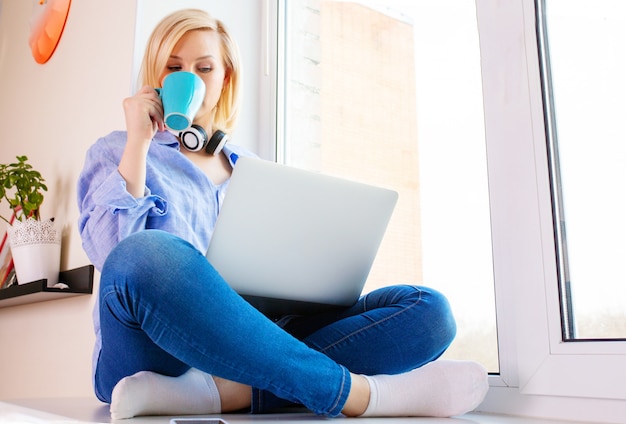 La jeune femme est assise sur le rebord de la fenêtre et travaille sur un ordinateur portable