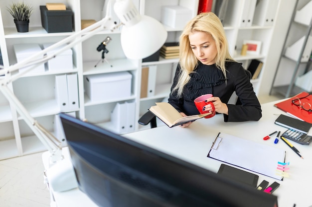 Une jeune femme est assise à une table dans le bureau, tenant une tasse et lisant un livre.