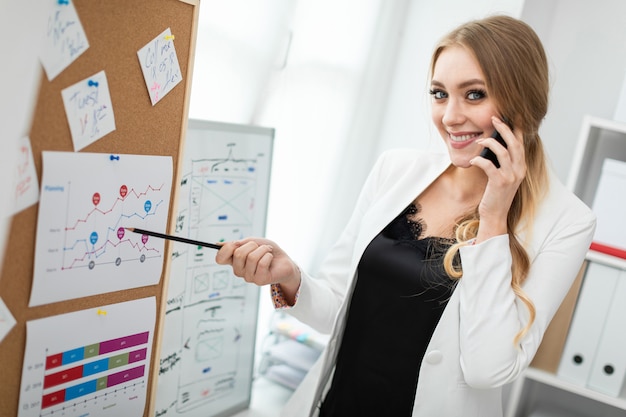 Une Jeune Femme Est Debout Près Du Tableau Avec Des Autocollants, Parle Au Téléphone Et Tient Un Crayon à La Main.