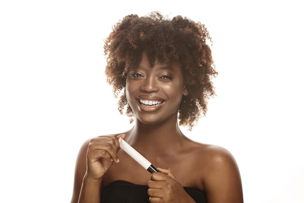 Jeune femme d'ethnie afro joyeuse avec une peau saine et propre tenant un tube d'hydratant sur un blanc