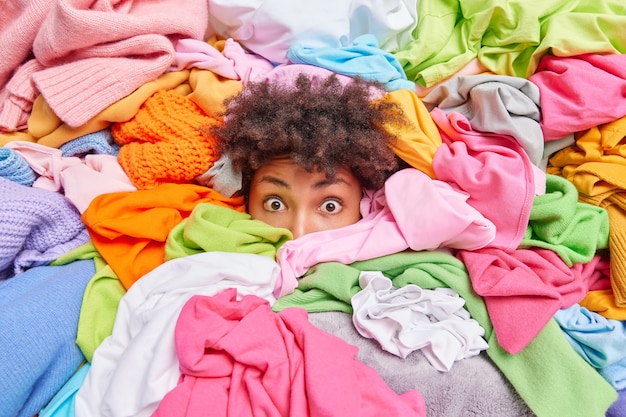 Une jeune femme ethnique aux cheveux bouclés surprise coincée dans un tas de linge fait des travaux ménagers pour plier des objets. Tête humaine dans un énorme tas de vêtements dépliés. Don de désencombrement et concept d'entretien ménager