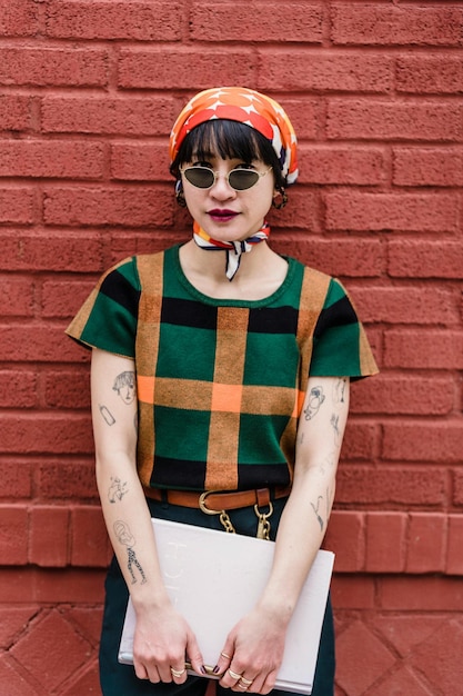 Une jeune femme ethnique cool en T-shirt à carreaux et lunettes de soleil avec un manuel regardant la caméra contre