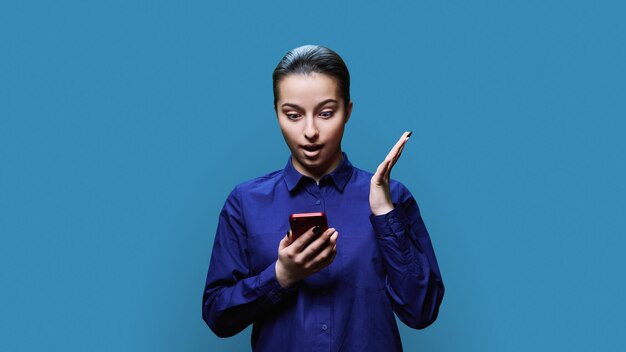 Une jeune femme étonnée, excitée et choquée avec un smartphone sur le fond bleu du studio Une adolescente utilisant des applications mobiles pour smartphone en train de regarder son téléphone