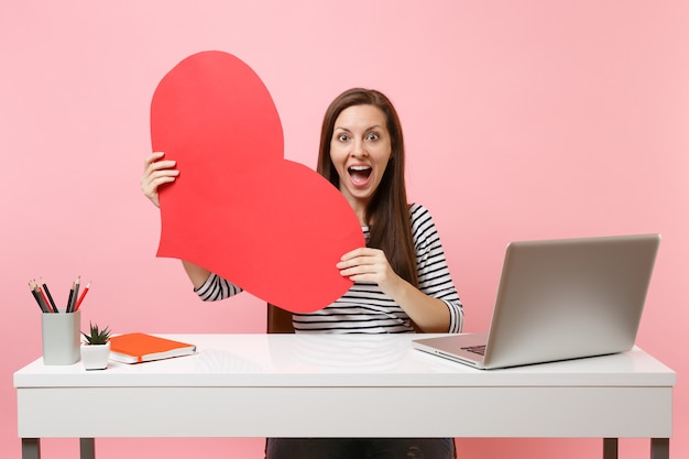 Jeune femme étonnée excitée tenant un coeur vide rouge s'asseoir, travailler au bureau blanc avec un ordinateur portable isolé sur fond rose pastel. Concept de carrière d'entreprise de réalisation. Copiez l'espace pour la publicité.