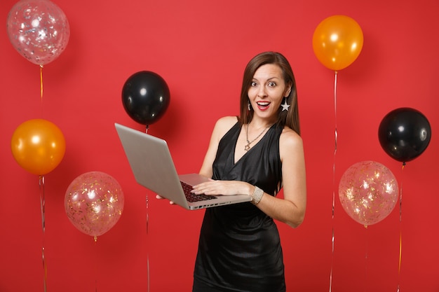 Jeune femme étonnée en petite robe noire travaillant sur un ordinateur portable tout en célébrant sur des montgolfières à fond rouge vif. Journée de la femme, bonne année, concept de fête de vacances de maquette d'anniversaire.