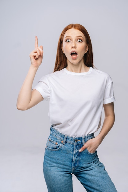 Jeune femme étonnée portant un t-shirt et un pantalon en jean pointant les doigts vers le haut avec la bouche ouverte