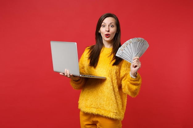 Jeune femme étonnée en pull de fourrure jaune tenant un ordinateur portable, fan d'argent en billets de banque en dollars, argent liquide isolé sur fond rouge. Émotions des gens, concept de style de vie. Maquette de l'espace de copie.