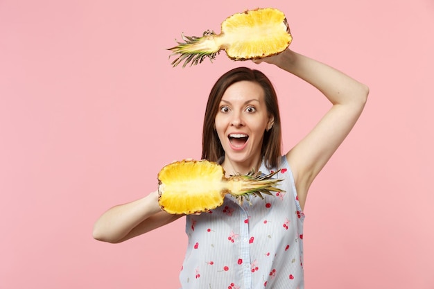 Une jeune femme étonnée en vêtements d'été gardant la bouche ouverte tient la moitié des fruits frais d'ananas mûrs isolés sur fond pastel rose. Les gens vivent un style de vie vivant et se détendent. Maquette de l'espace de copie.