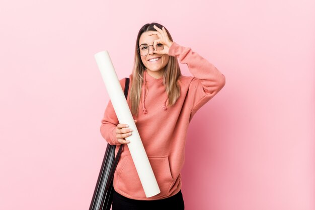 Jeune femme étudiante en architecture excitée en gardant le geste ok sur les yeux.