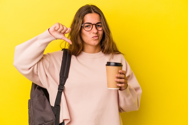 Jeune femme étudiante caucasienne tenant un café isolé sur fond jaune montrant un geste d'aversion, les pouces vers le bas. Notion de désaccord.