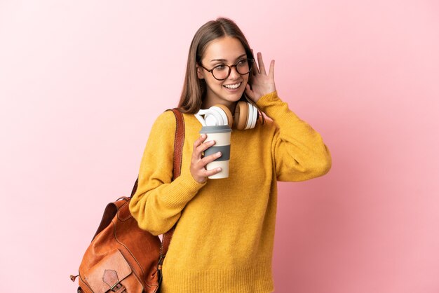 Jeune femme étudiante sur un mur rose isolé à l'écoute de quelque chose en mettant la main sur l'oreille