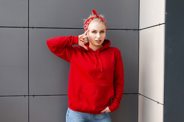 Jeune femme européenne avec une coiffure à la mode avec un bandana rouge à la mode dans un sweat-shirt rouge élégant avec une capuche en jeans pose près du bâtiment moderne gris sur une chaude journée de printemps