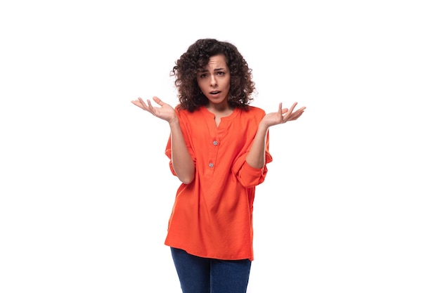 Une jeune femme européenne confuse avec des cheveux noirs bouclés dans une chemise orange se sent surprise