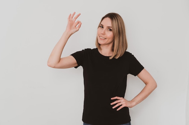 Jeune femme européenne dans des vêtements décontractés pointant avec le doigt ok chanter sur fond blanc. Gestes de la main