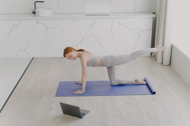 Jeune femme européenne heureuse fait de l'exercice de planche et regarde dans un ordinateur portable Entraînement en ligne à la maison
