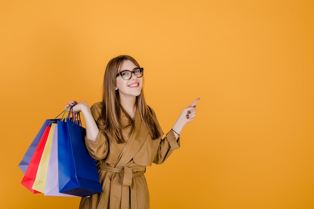 Jeune femme européenne hipster dans des verres et manteau avec des sacs à provisions colorés isolés sur jaune