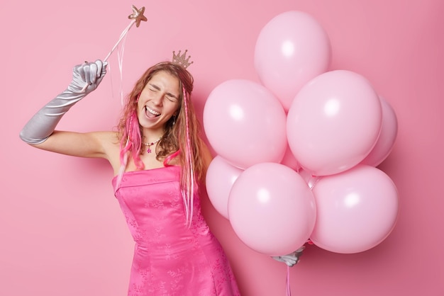 Une jeune femme européenne insouciante porte une robe et des gants, danse et rit positivement, l'ambiance festive vient à la fête célèbre quelque chose qui contient un tas de ballons à l'hélium isolés sur fond rose