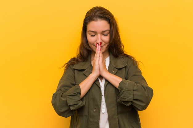 Jeune femme européenne isolée sur jaune se tenant la main en priant près de la bouche, se sent confiant.