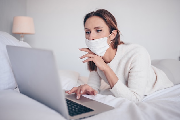 Jeune femme européenne en masque de médecine du visage travaillant sur un ordinateur portable dans la chambre pendant l'isolement du coronavirus