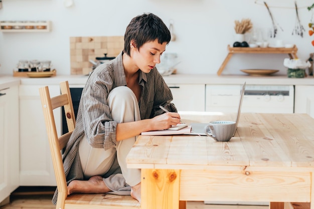 Une jeune femme européenne moderne aux cheveux courts travaille à la maison sur un ordinateur assise à une table en bois.