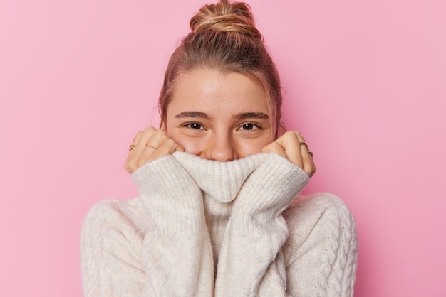 Une jeune femme européenne positive aux cheveux blonds rassemblés en chignon porte un pull blanc chaud tricoté par temps froid couvre la bouche avec un col semble heureusement isolée sur fond rose