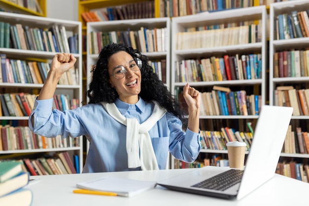 Photo une jeune femme excitée dans une bibliothèque lève ses poings en signe de victoire avec un ordinateur portable et une tasse de café sur le