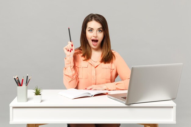 Jeune femme excitée dans des vêtements décontractés pastel écrivant des notes, s'asseoir et travailler au bureau blanc avec ordinateur portable pc isolé sur fond gris. Concept de style de vie de carrière d'affaires de réalisation. Maquette de l'espace de copie.