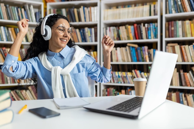 Photo une jeune femme excitée avec des écouteurs dansant joyeusement à un bureau de bibliothèque entourée de livres.