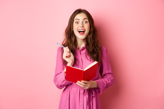 Une jeune femme excitée a une excellente idée, écrivant ses idées dans un planificateur, tenant un carnet de notes et souriant étonné, debout sur un mur rose.