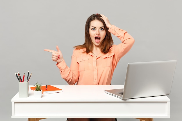 Jeune femme excitée mettant la main sur la tête, pointant l'index de côté, asseyez-vous au bureau avec un ordinateur portable pc isolé sur fond gris. Concept de style de vie de carrière d'affaires de réalisation. Maquette de l'espace de copie.