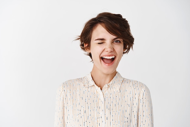 Jeune femme excitée et motivée faisant un clin d'œil heureux souriant et ressentant de la joie, debout sur un mur blanc