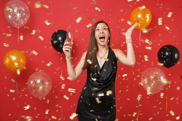 Jeune femme excitée en petite robe noire célébrant, tenant une coupe de champagne et attrapant des confettis sur des montgolfières à fond rouge vif. Bonne année, concept de fête de vacances de maquette d'anniversaire.