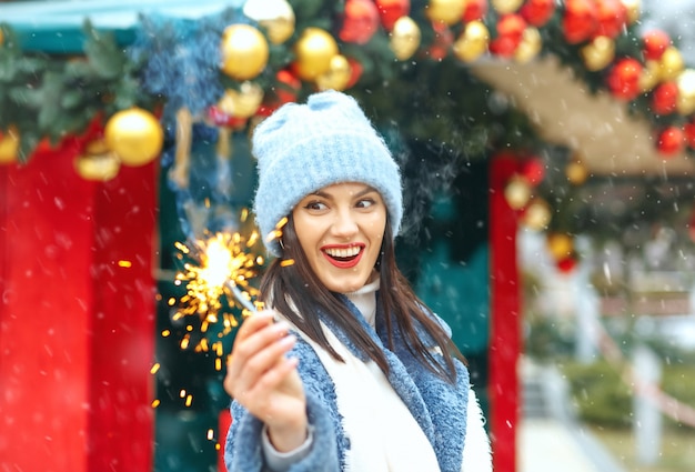 Une jeune femme excitée porte un manteau bleu profitant de vacances avec des lumières du Bengale pendant les chutes de neige