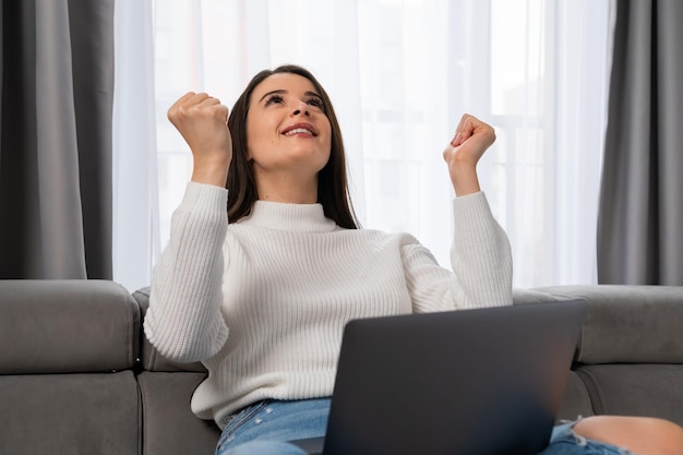 Une jeune femme excitée se réjouit de la victoire sur Internet tenant un ordinateur portable sur ses genoux dans un salon confortable