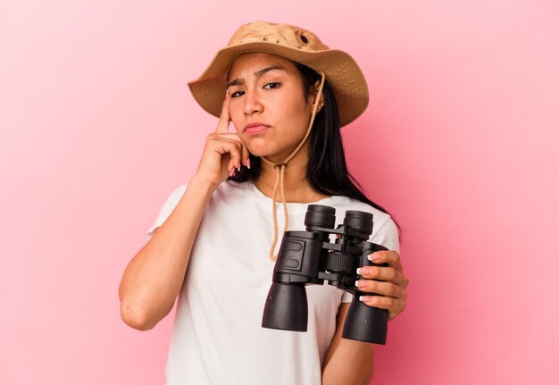 Jeune femme exploratrice de race mixte tenant des jumelles isolées sur fond rose pointant le temple avec la pensée du doigt concentrée sur une tâche