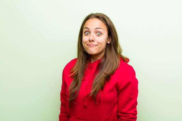 Jeune femme avec une expression maladroite, folle et surprise, les joues gonflées, se sentant bourré, gras et plein de nourriture contre le mur vert