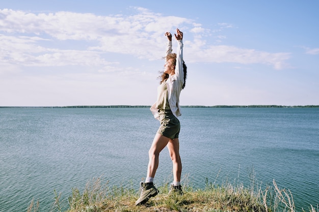 Jeune femme extatique avec ses bras levés en gardant les yeux fermés en se tenant debout sur la berge par l'eau et profitant de la solitude