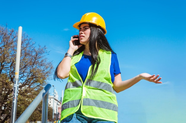 Une jeune femme à l'extérieur portant un casque et un gilet de sécurité jaune discute du téléphone avec son patron