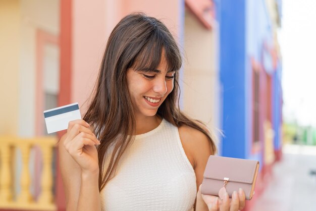 Jeune femme à l'extérieur tenant un portefeuille et une carte de crédit avec une expression heureuse