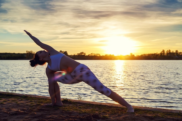 Jeune femme faisant des exercices de yoga sur la plage du lac au coucher du soleil