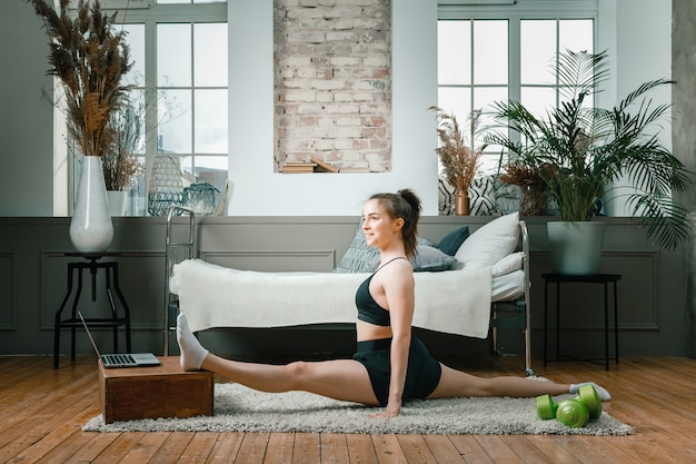 Une jeune femme fait du sport à la maison, entraînement en ligne. L'athlète s'étire, médite, assis sur une ficelle dans la chambre, à l'arrière-plan il y a un lit, un vase, un tapis.