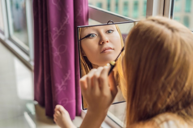 La jeune femme fait le maquillage s'asseyant par la fenêtre avec une vue panoramique des gratte-ciel et du grand