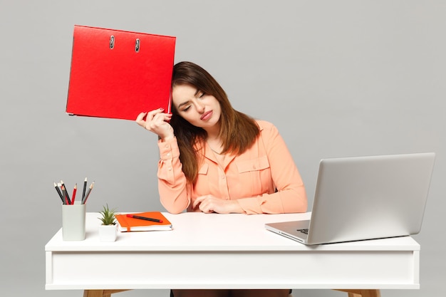 Jeune femme fatiguée dans des vêtements pastel tenant un dossier rouge pour document papiers, travail au bureau avec ordinateur portable pc isolé sur fond gris. Concept de style de vie de carrière d'affaires de réalisation. Maquette de l'espace de copie.