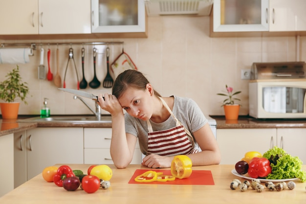 La jeune femme fatiguée émaciée dans un tablier cuisine dans la cuisine à la maison. Concept de régime. Mode de vie sain. Préparer la nourriture.