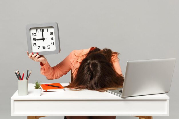 Jeune femme fatiguée épuisée avec la tête baissée tenant une horloge carrée, s'asseoir et travailler au bureau avec un ordinateur portable pc isolé sur fond gris. Concept de style de vie de carrière d'affaires de réalisation. Maquette de l'espace de copie.