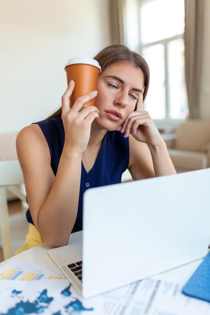 Jeune femme fatiguée regardant loin et tenant une tasse de café près de la tête au bureau sur un premier plan flou