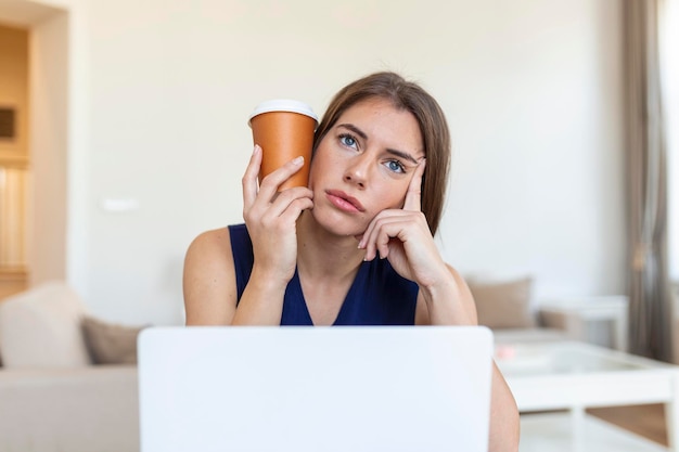 Jeune femme fatiguée regardant loin et tenant une tasse de café près de la tête au bureau sur un premier plan flou