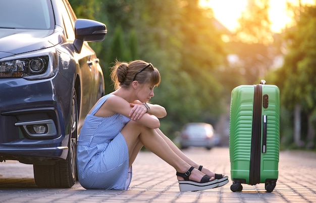 Jeune femme fatiguée avec une valise assise à côté de la voiture en attente de quelqu'un. Concept de voyage et de vacances.