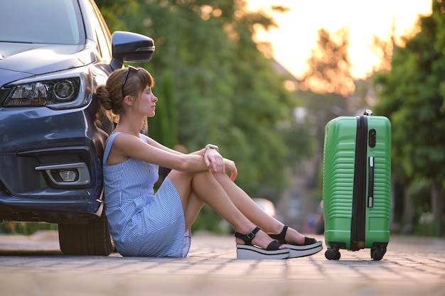 Jeune femme fatiguée avec une valise assise près de sa voiture en attente de quelqu'un. Concept de voyage et de vacances.