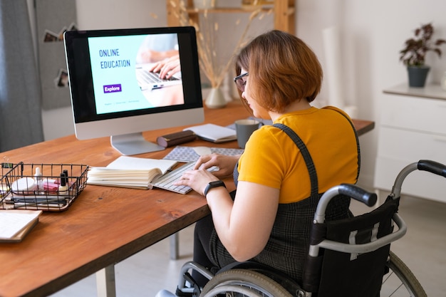 Jeune femme en fauteuil roulant en appuyant sur les touches du clavier en face de l'écran de l'ordinateur tout en allant avoir une leçon à distance dans l'environnement familial